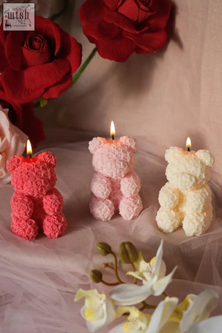 Three lit teddy rose-shaped candles in pink and cream hues shaped like small teddy bears with rose texture