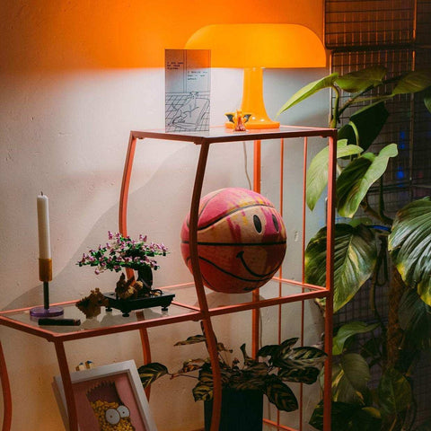 A vibrant pink and orange basketball with a smiling face sits on an orange shelf near the ambient mushroom desk lamp