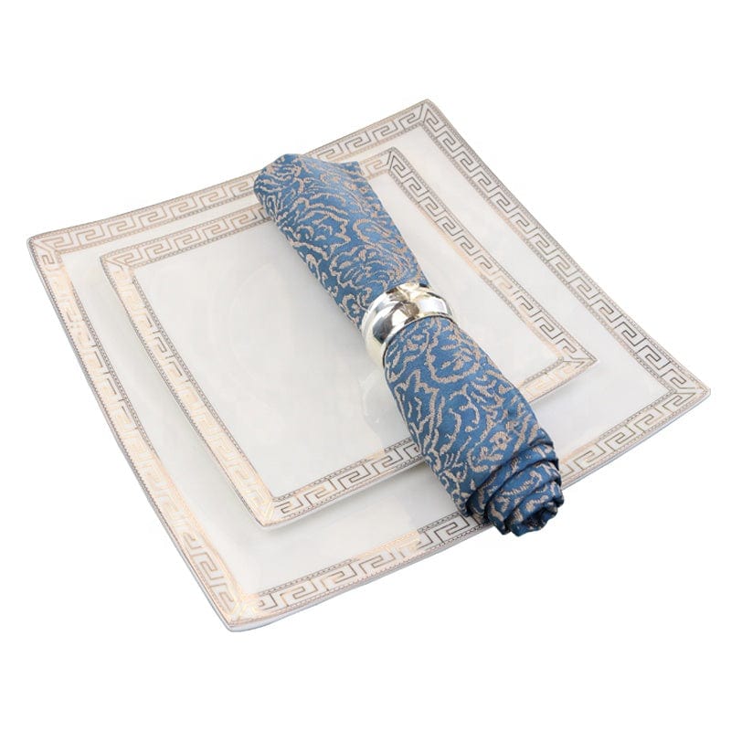 The glamour bone china dinner set features white square plates with greek key border and a blue patterned napkin rolled with a silver ring