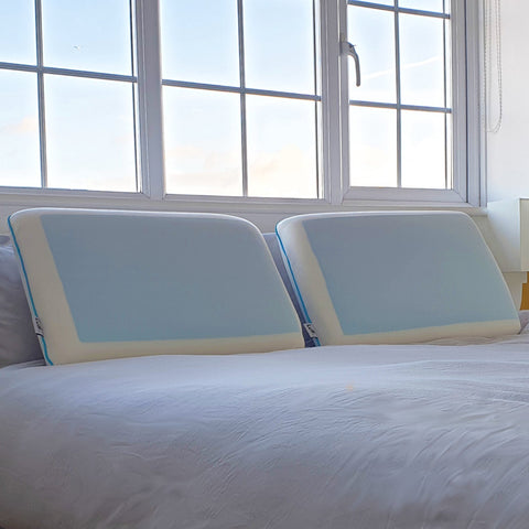 Two light blue memory foam pillows with white edges and teal trim for cooling comfort near a window bed