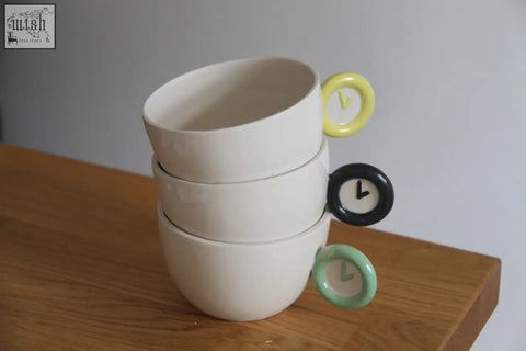 Stack of three white ceramic time zone cups with colorful clock face handles in yellow black and mint green