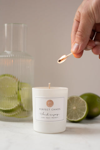 White cylindrical travel candle with minimalist perfect chaos label and circular logo set against lime slices