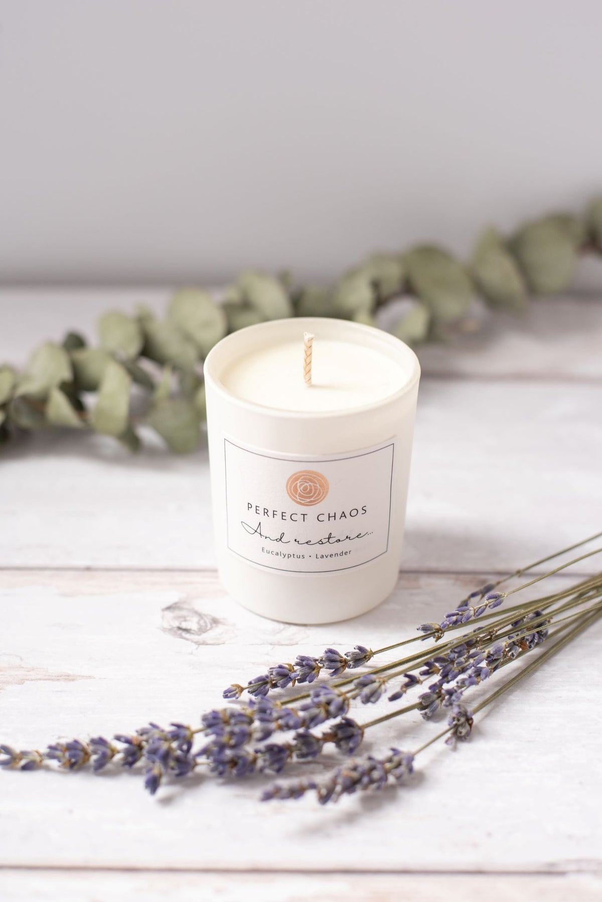 A white cylindrical travel candle with perfect chaos and restore label featuring eucalyptus lavender essential oils scent