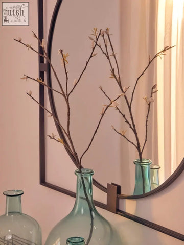 A slender teal glass vase with brown branches inside a large square black metal frame round insert wall mirror