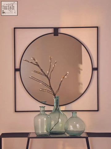 Large square black metal frame round insert wall mirror with frosted glass center above green glass vases