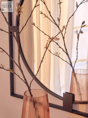 Tall amber glass vase with brown budding branches next to a small square black metal frame wall mirror
