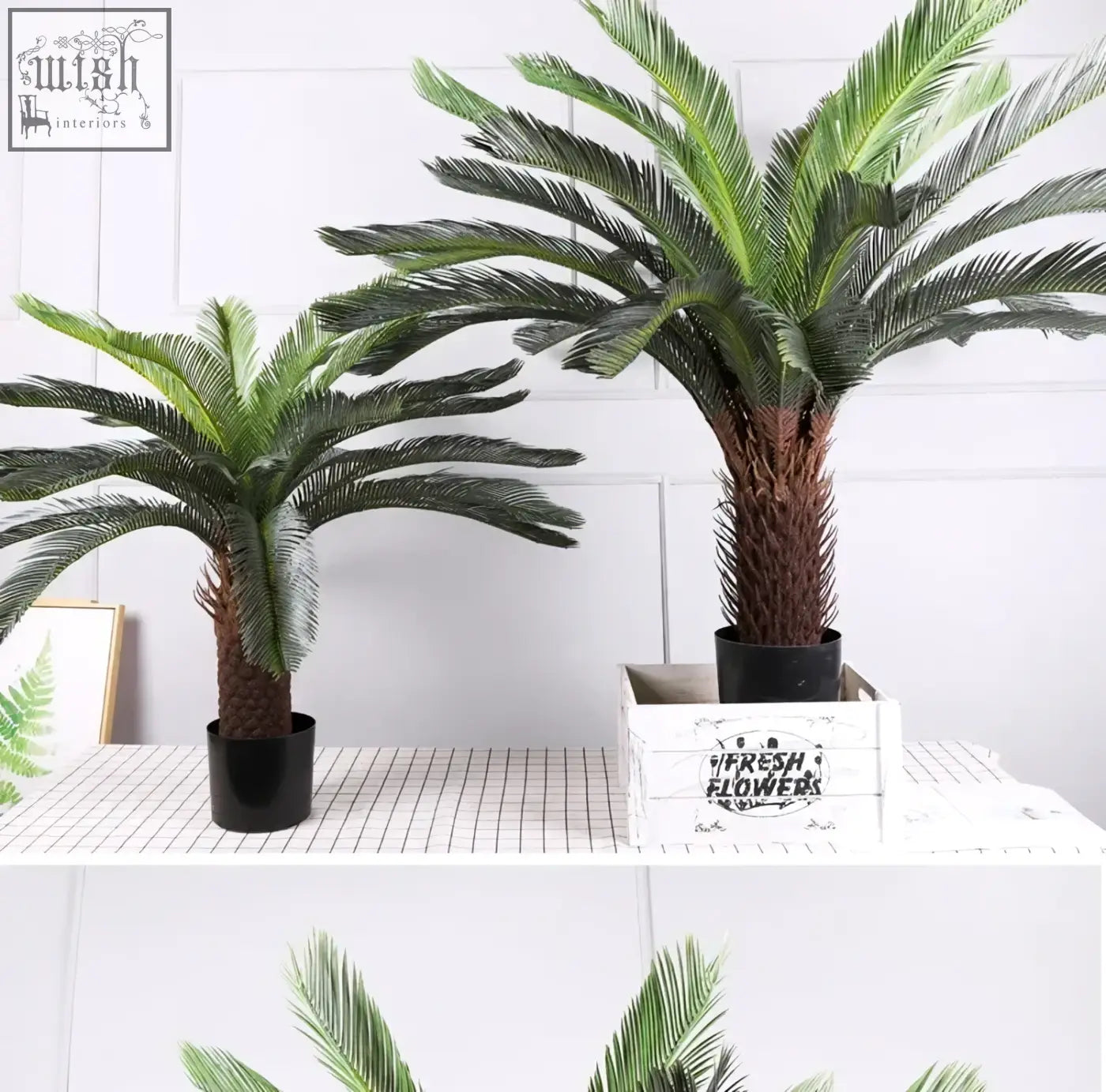 Two tropical artificial palm trees with dark green plastic palm leaves and textured brown trunks in black pots for decor