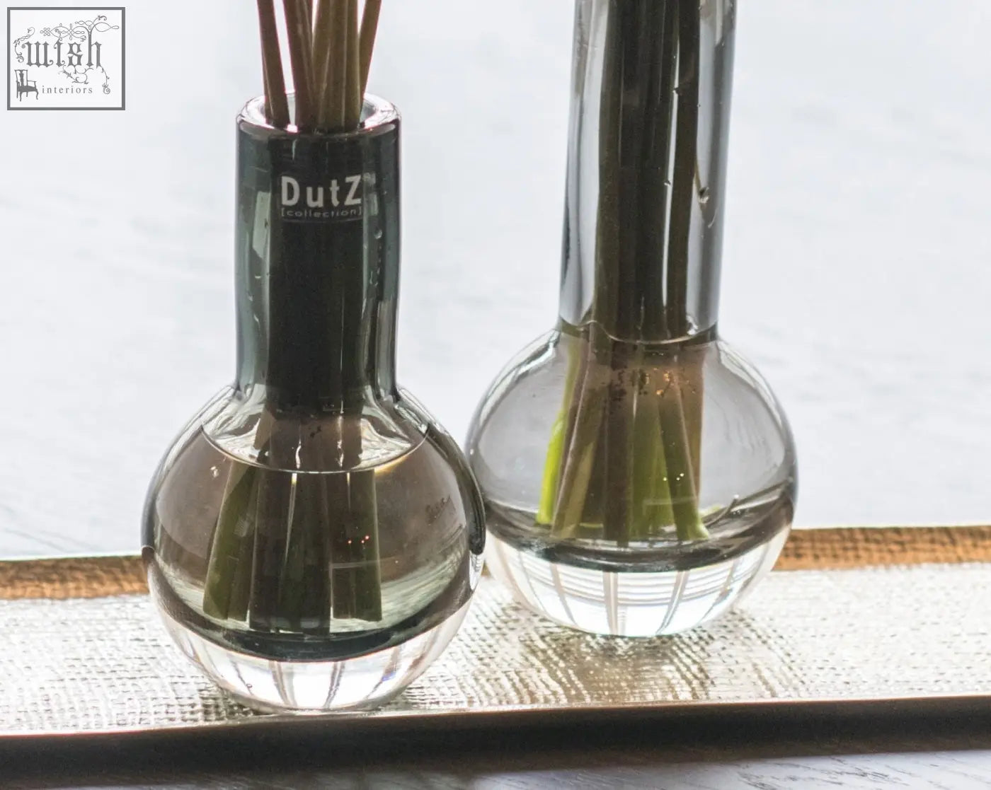 Two clear mouth blown glass reed diffusers with black caps from dutz collection holding brown reeds in vase masa grey