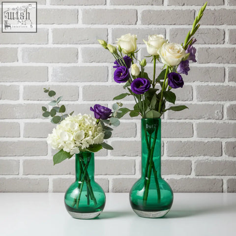 Two emerald green mouth blown glass vases from the dutz collection holding white roses purple lisianthus and hydrangeas
