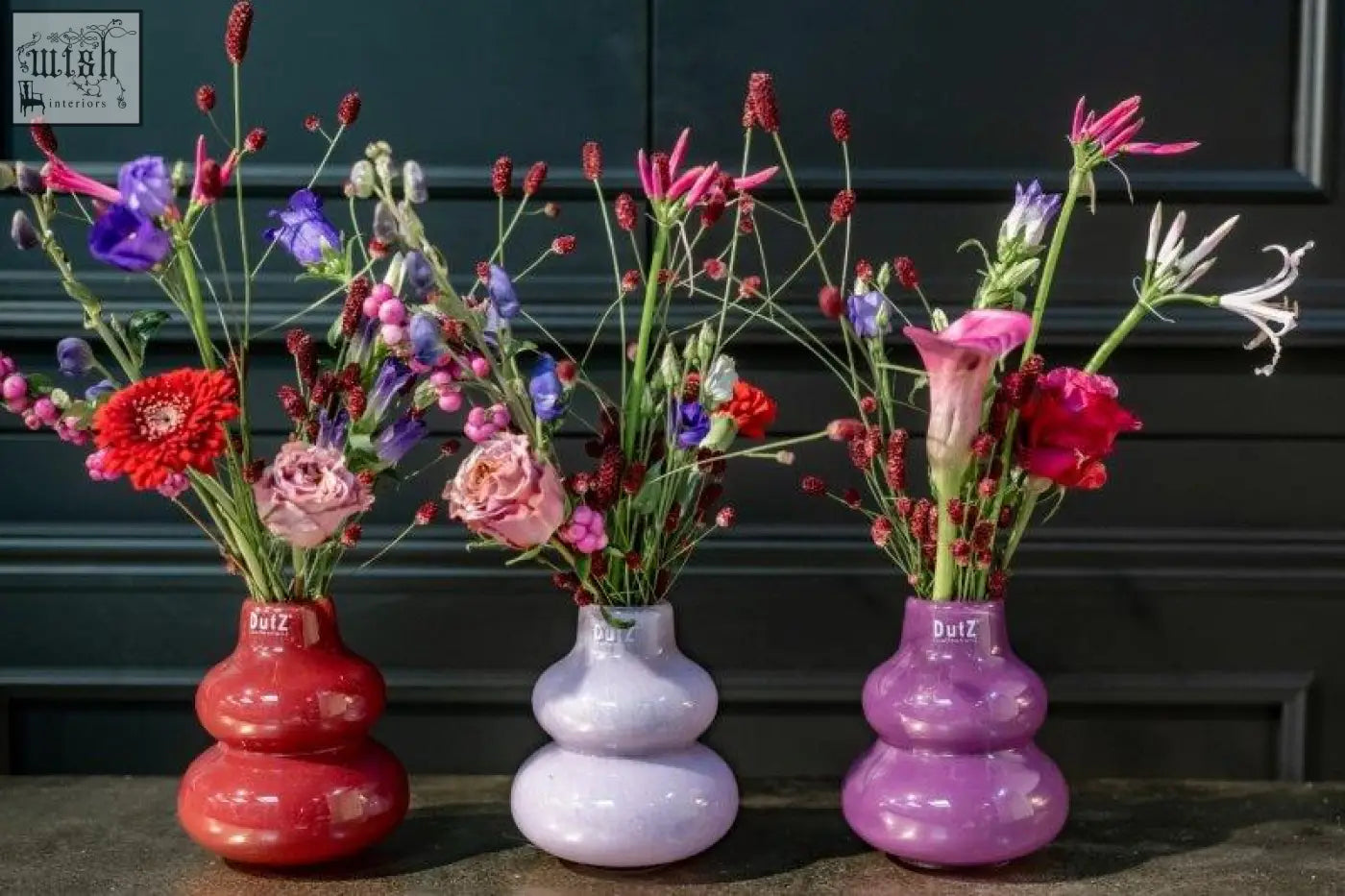 Three colorful mouth blown dutz collection vases in red lavender and purple holding vibrant mixed flowers