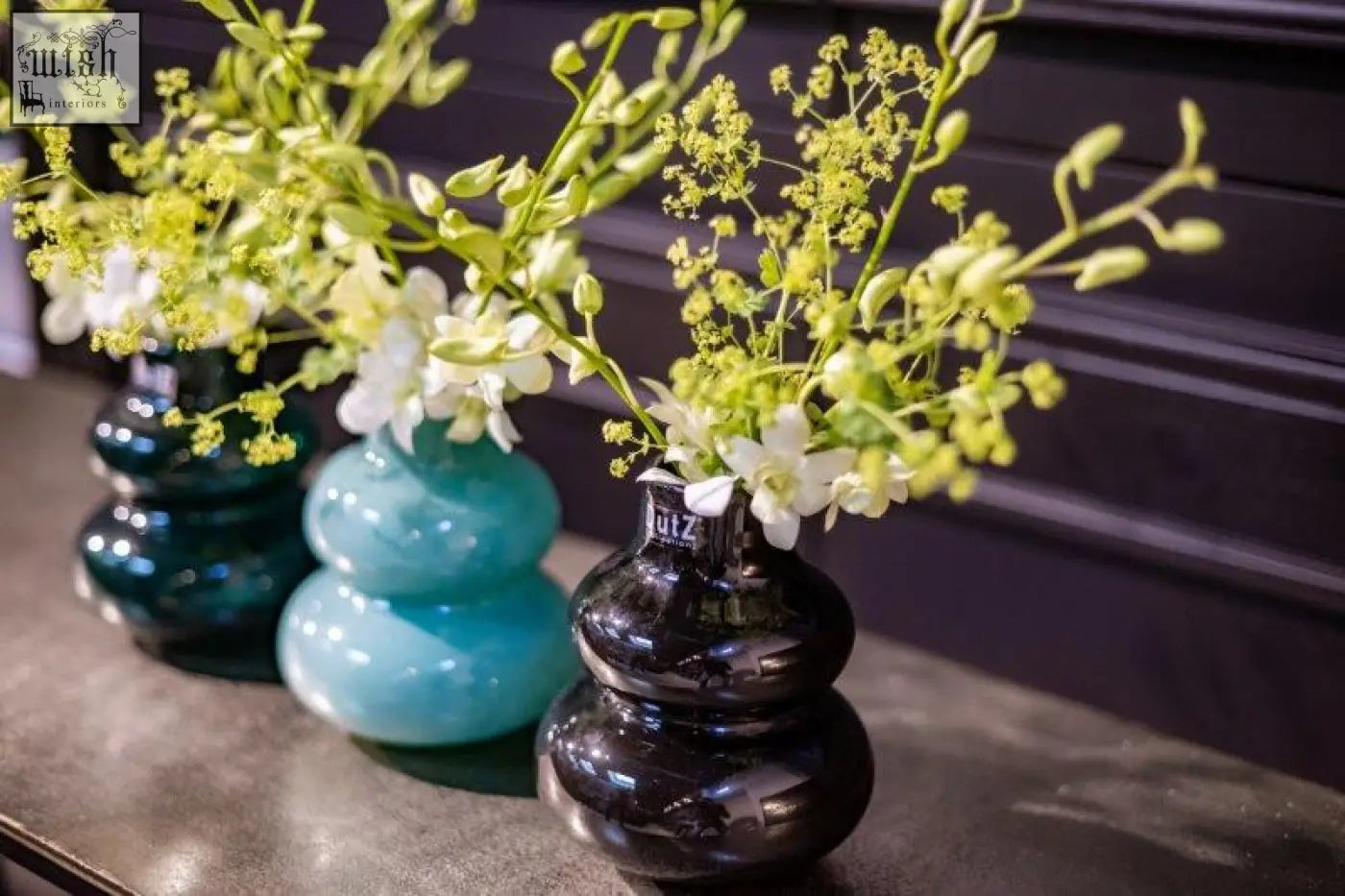 Three mouth blown glass vases from dutz collection in teal and black hold white orchids with green buds