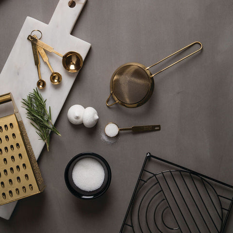Gold measuring spoons with polished finish resting on a white marble cutting board near vertex black nickel square trivet