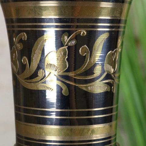 Black and gold engraved metal tumbler with floral patterns complementing a large brass vase with silver and gold polish