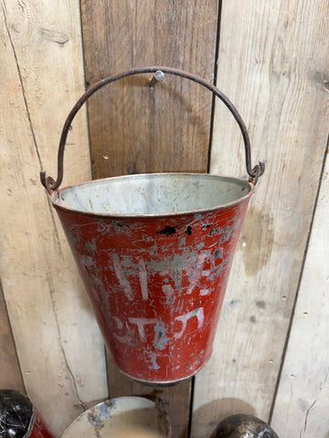 A weathered red vintage galvanised metal hanging fire bucket with rusted wire handle against wooden wall