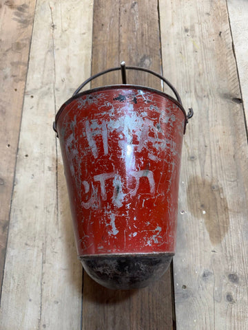 Vintage red enamel hanging fire bucket with metal handle and faded white lettering mounted on wooden surface
