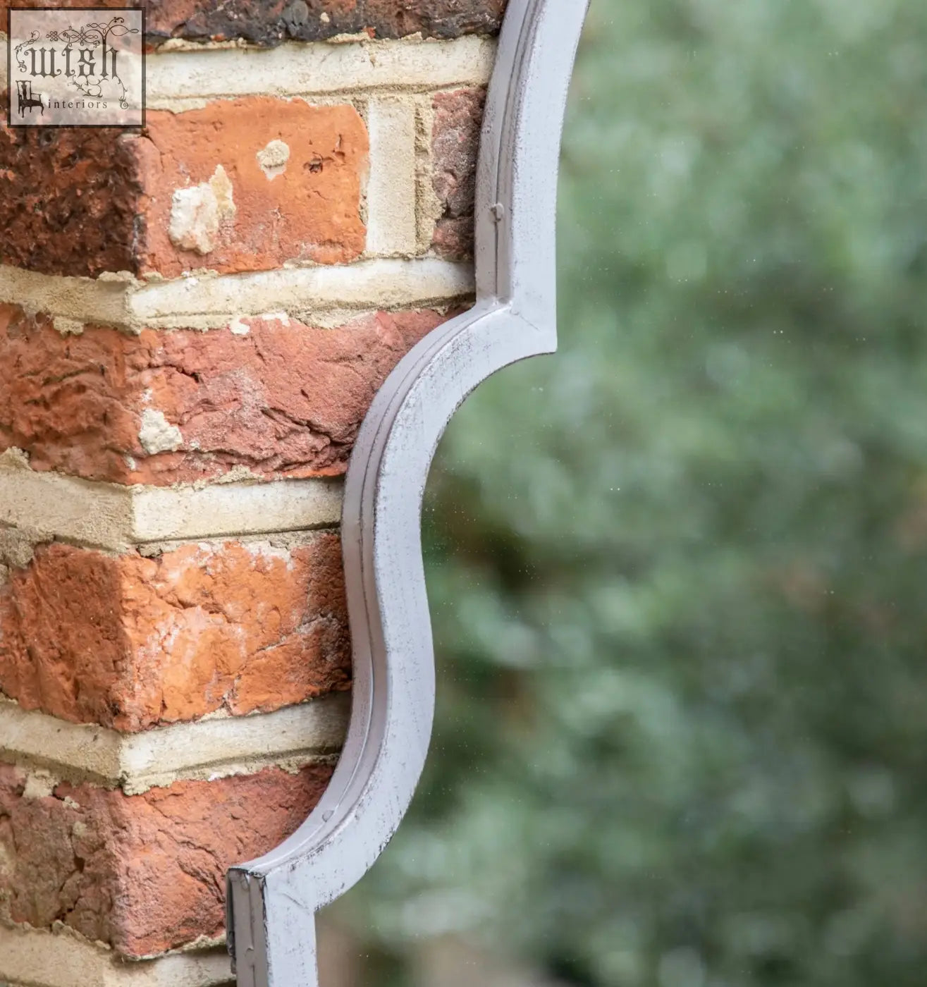 Vintage grey metal outdoor mirror with a curved ornate edge leaning against a red brick wall