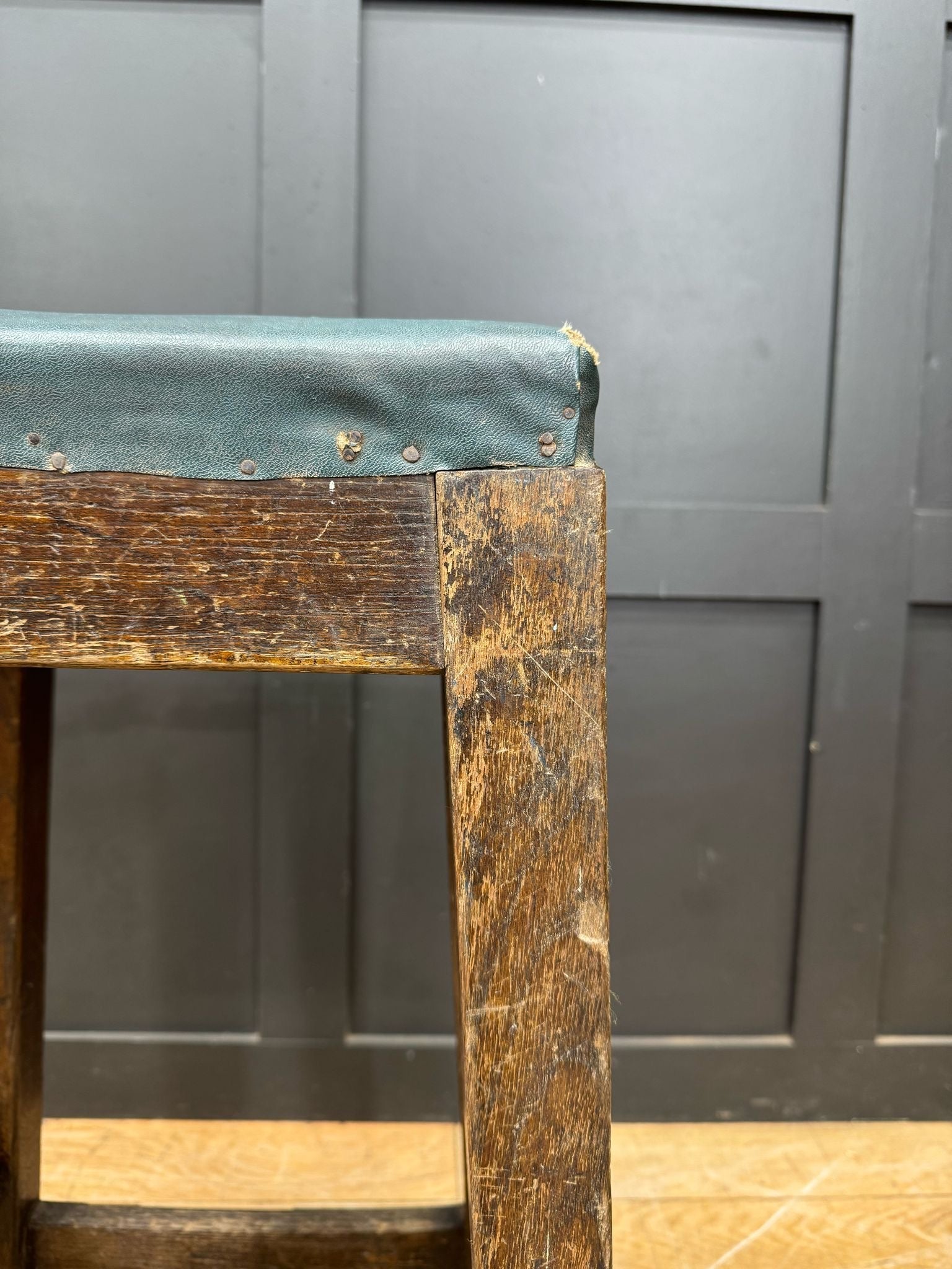 A vintage railway stool with a worn wooden frame and teal leather seat reinforced by metal studs along the edge
