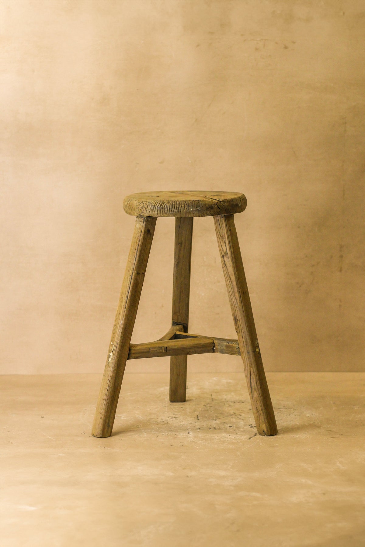 Vintage rustic elm wood stool with round weathered seat and four splayed legs joined by a central crossbar