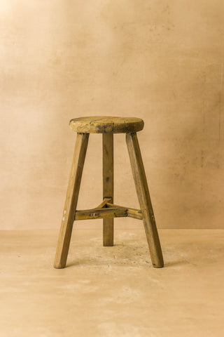 Vintage rustic elm wood stool with a round weathered seat and three angled legs joined by a central crossbar wood stool