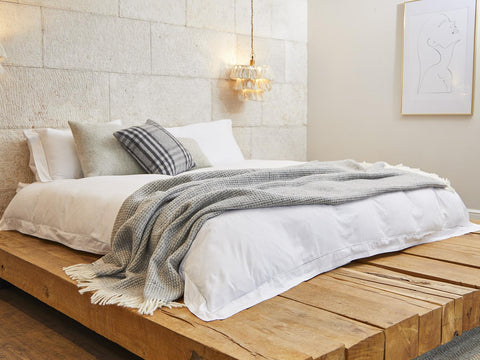A cozy silver grey waffle xl pure new wool throw blanket with frayed edges draped over white bedding on a wooden bed