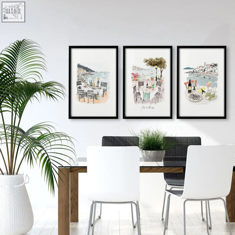 Three black-framed watercolor prints of mediterranean coastal dining scenes as wall art for a dining room above a wooden table