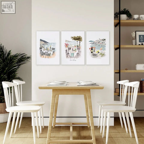 Light wood dining table with slender legs and minimalist design next to mediterranean coastal wall art set of three prints