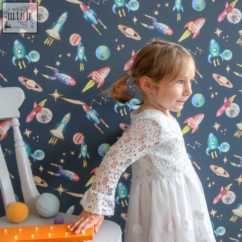 A young girl in a white lace dress stands against dark blue hand-painted space wallpaper with rockets and planets