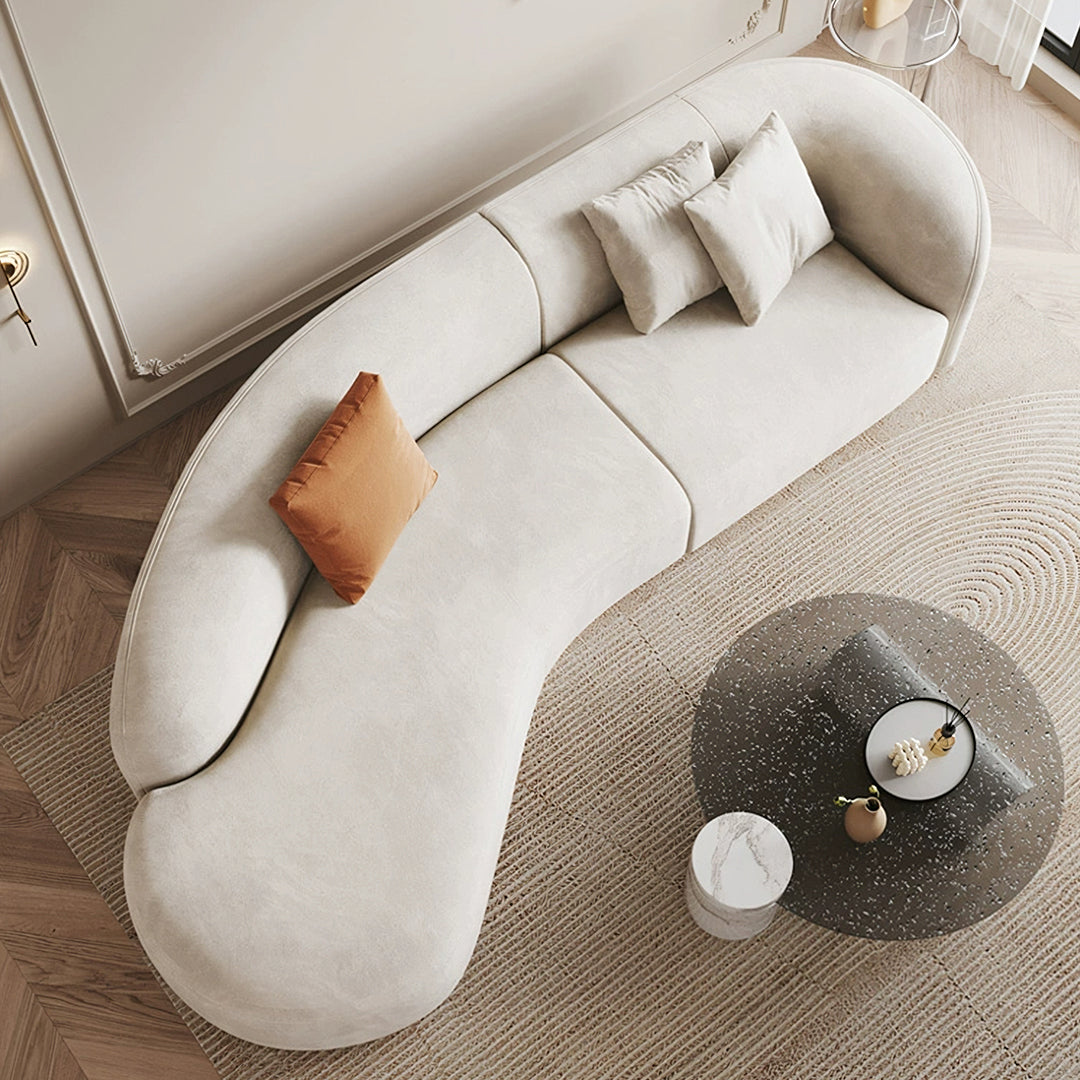 Cream-colored welby three seater curved sofa with soft velvet upholstery and a rust-orange throw pillow