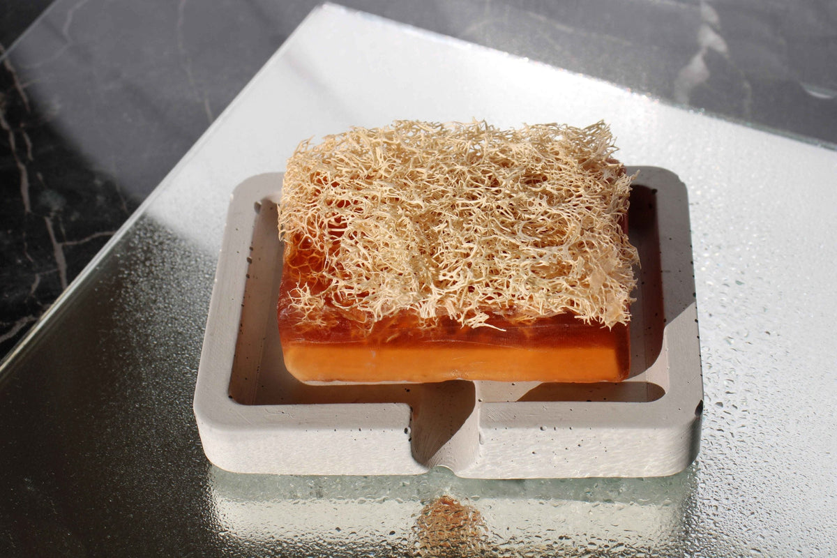 A rectangular orange soap with beige loofah sits in a minimalist white concrete soap holder for bathroom use
