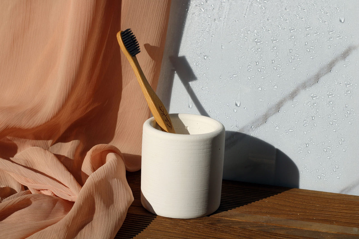 A natural wood toothbrush with black bristles in a white concrete toothbrush holder cup with minimalist design