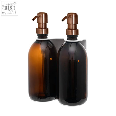 Pair of amber glass bottles with copper pumps in a white wall-mounted double soap dispenser unit