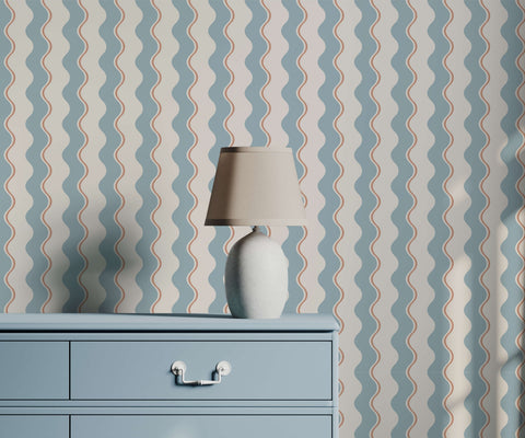 A sleek off-white ceramic wiggle table lamp with a tapered shade on a light blue dresser against wavy stripe wallpaper