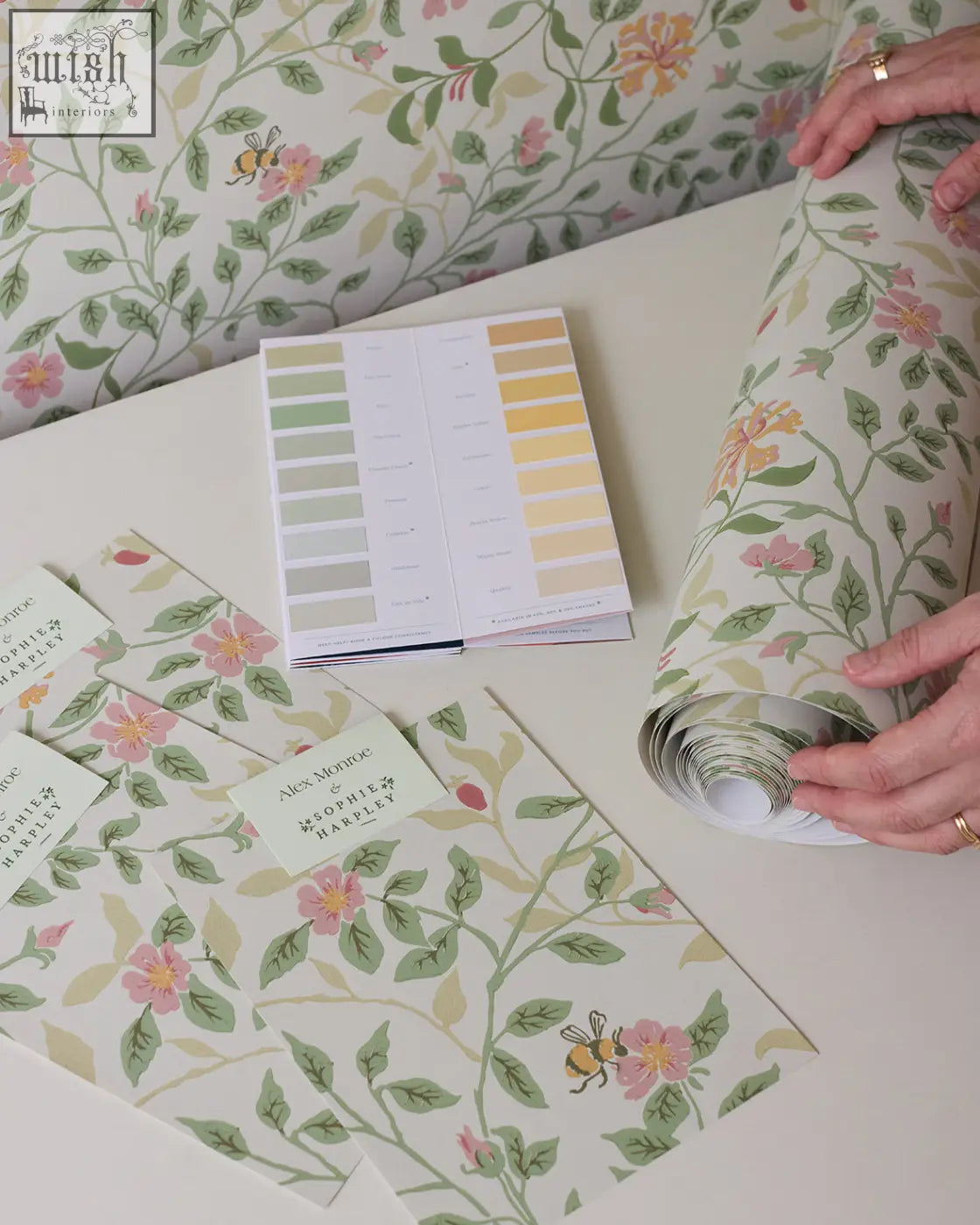 A roll of wild rose wallpaper by alex monroe featuring soft pink roses green leaves and bees on an off-white background