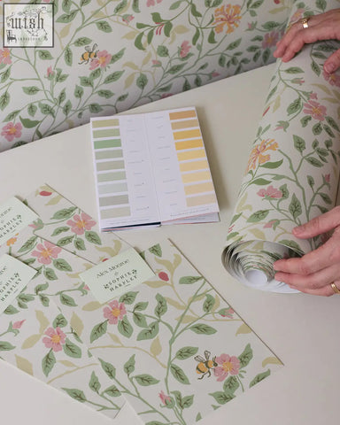 A roll of wild rose wallpaper by alex monroe featuring soft pink roses green leaves and bees on an off-white background