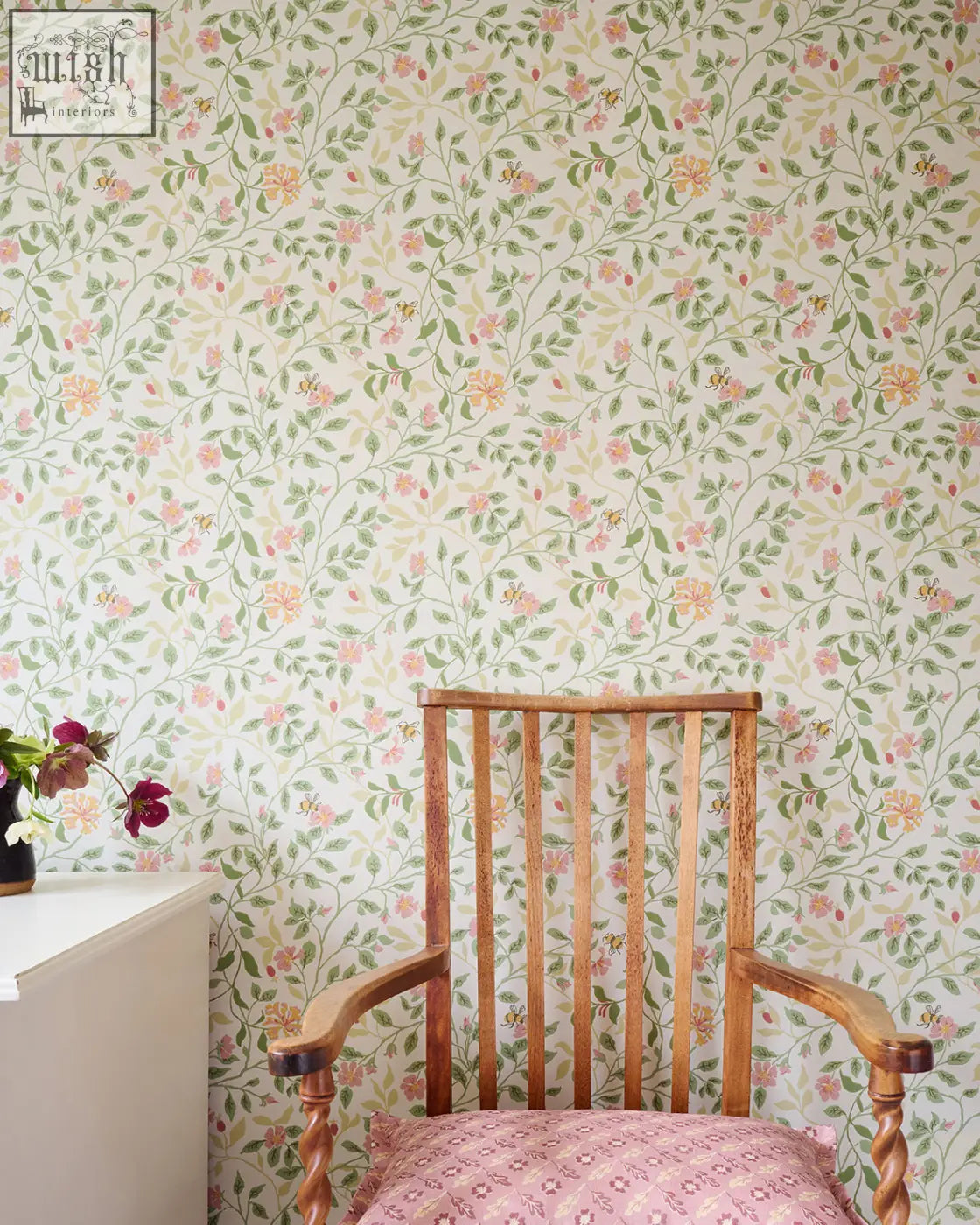 A wooden rocking chair with a pink patterned cushion against alex monroe wild rose wallpaper in peridot green