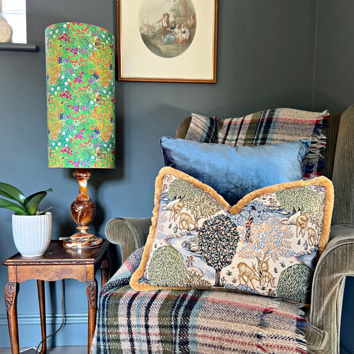 A william morris deer garden tapestry cushion with mustard gold 30mm fringe sits by a green floral lampshade on wood table