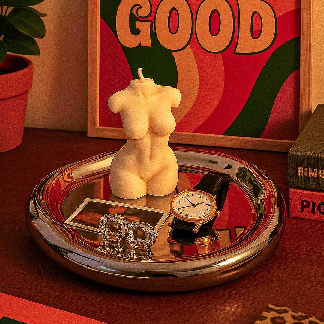 Cream-colored sculpted female torso candle with wick on a shiny wobbly chrome tray for stylish decor