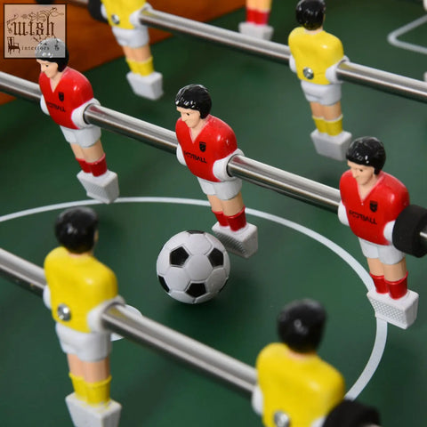 Wooden football table with red and yellow foosball players and a black and white soccer ball on green playing field