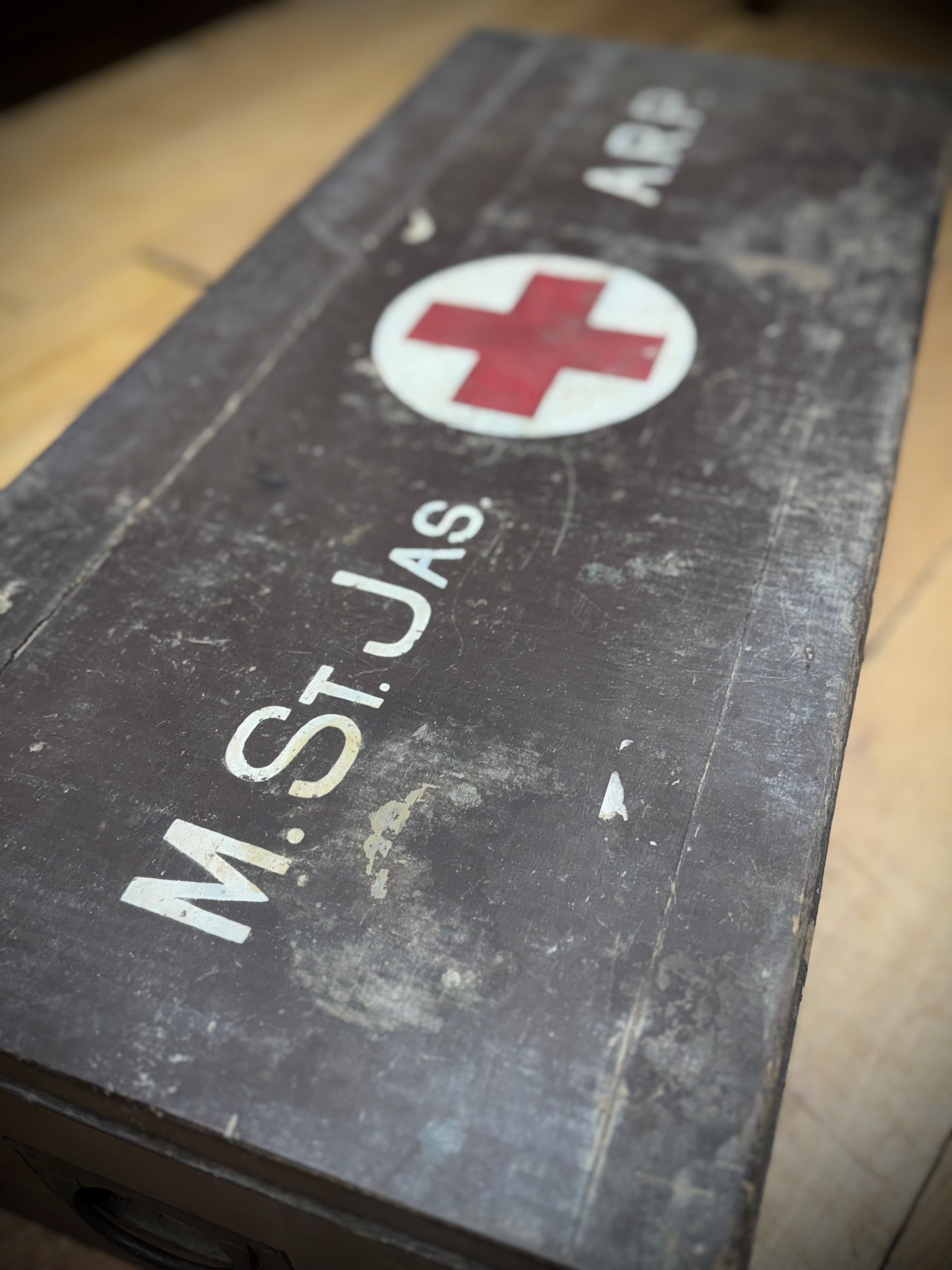 A weathered dark gray metal wwii civil defence aid box with red cross emblem and white lettering for medical use