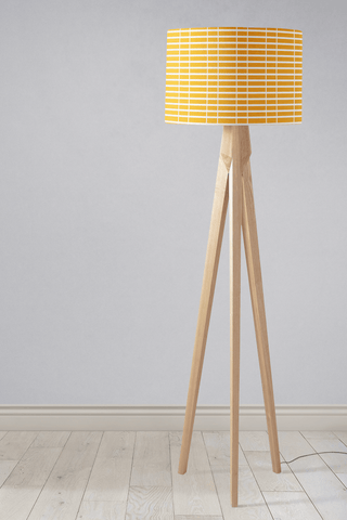 A modern tripod table lamp with a yellow lamp shade featuring white geometric stripes and light wooden legs