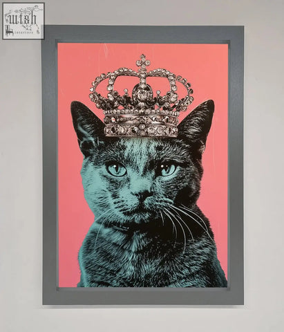 A framed print of a majesty cat framed featuring a black cat with teal eyes and a jeweled silver crown on pink background