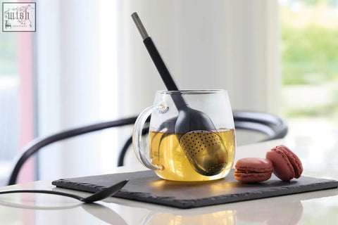 Clear glass mug with handle holding golden tea and a black perforated stainless steel tea ball stick infuser spoon