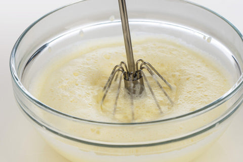 A stainless steel immersion blender with spin whisks from zamin set mixing pale yellow batter in a glass bowl