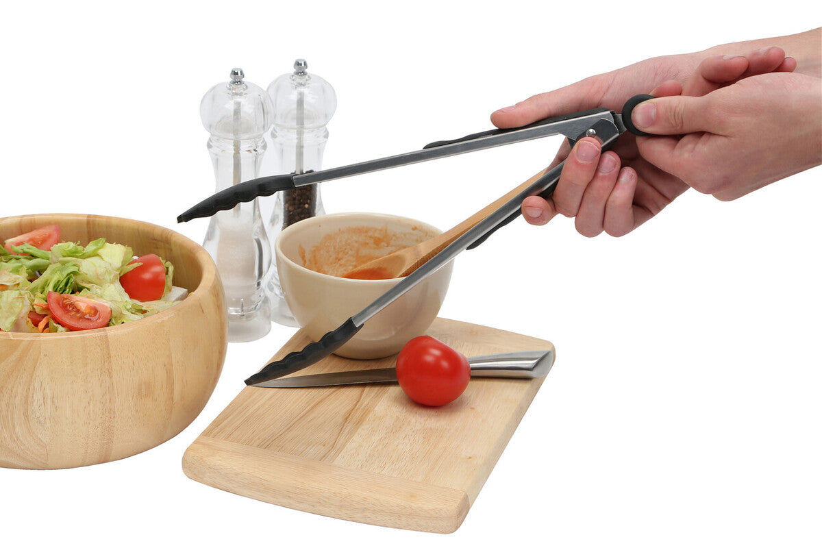 Silver-toned stainless steel tongs with black silicone handles gripping a tomato on a wooden cutting board